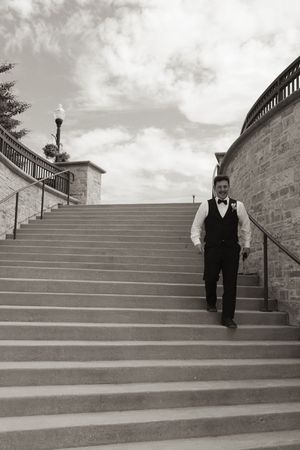 Groom in black tuxedo vest and bow tie walks down grand stone staircase in black and white photo.