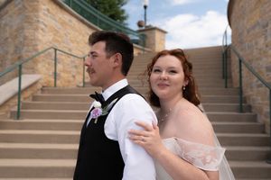 A bride and groom pose back-to-back on stone steps, the bride in an off-shoulder gown and groom in a black vest.