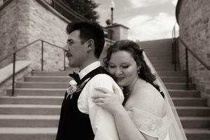 Black and white photo of bride and groom posing on stone steps, bride embracing groom from behind in wedding attire.