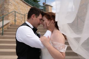 Bride and groom share an intimate forehead-to-forehead moment, with wedding veil billowing in the breeze on stone steps.