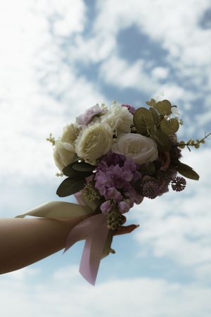 Bride holding a beautiful wedding bouquet of white roses and purple flowers against a cloudy blue sky.