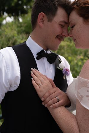 Bride and groom touching foreheads tenderly, showing wedding rings, groom in black vest and bow tie with floral boutonniere.