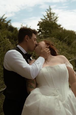 A bride and groom share a romantic kiss outdoors surrounded by green trees on their wedding day.