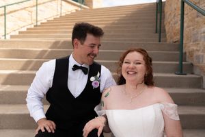 Happy newlywed couple sitting on stone steps, groom in black vest and bride in off-shoulder gown.