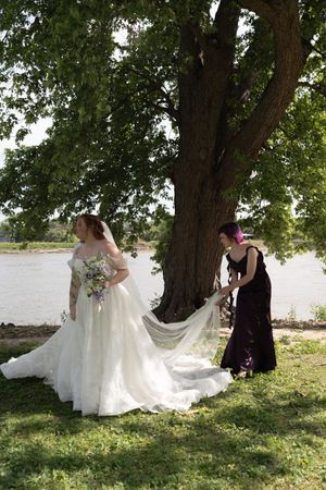 Bride in white ball gown holds bouquet while bridesmaid adjusts her veil under a large tree by a river.