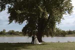 Bride and groom standing under a large tree by a river on their wedding day.