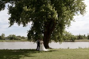 Bride and groom share a romantic moment under a large tree by a serene lake during their outdoor wedding ceremony.