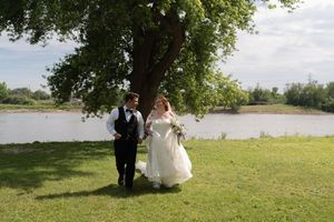 Bride and groom walking hand in hand on green lawn beside a serene river under a tree.