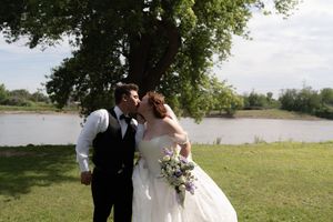 Bride and groom sharing a kiss near a tree by a lake, bride holding purple and white bouquet.