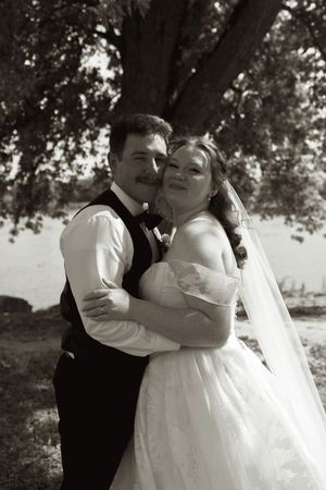 Black and white wedding portrait of smiling bride and groom embracing under a large tree outdoors.