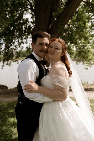 Bride and groom embrace outdoors near a large tree by a lake on their wedding day.