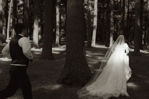 Black and white photo of bride and groom walking through a sunlit forest, bride's veil trailing behind her.