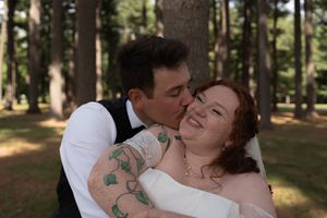 A groom with tattoos kisses his smiling red-haired bride on the cheek in a forest setting.