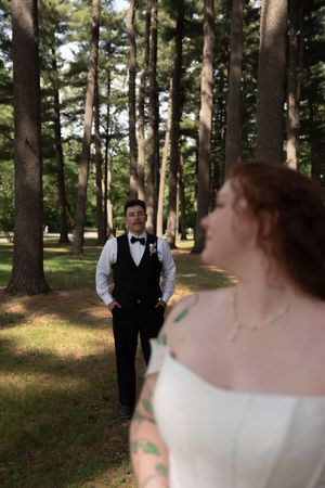Groom in black vest and bow tie stands among tall trees as bride in white dress poses in foreground.