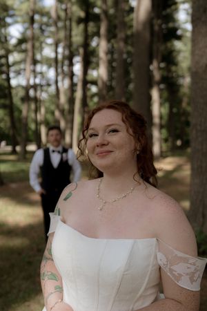 Red-haired bride in off-shoulder white gown smiles outdoors with groom standing among tall trees behind her.