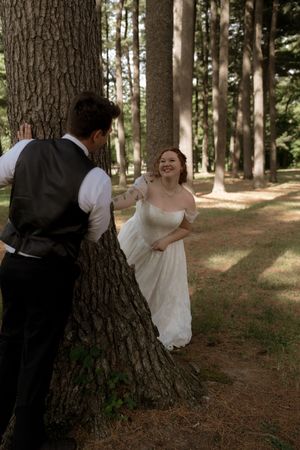 Bride in white gown smiles at groom leaning against a pine tree during a playful outdoor wedding photo session.