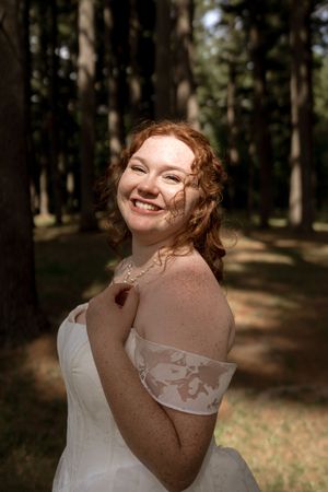 Smiling red-haired bride in an off-shoulder lace wedding dress posing in a sunlit forest.