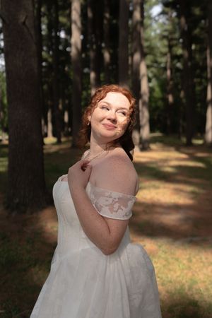 Radiant bride in white off-shoulder lace gown posing among tall pine trees in sunlit forest.