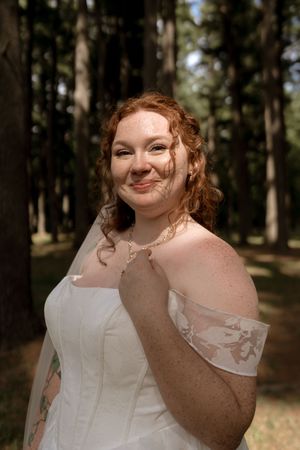 Smiling red-haired bride with freckles and tattoos poses in white off-shoulder wedding gown among tall trees.