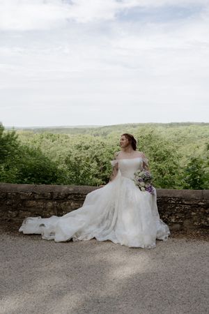 Elegant bride in off-shoulder ball gown wedding dress sitting on stone wall with lush green landscape backdrop.