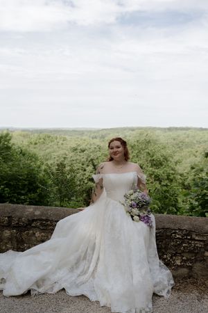 Bride in off-shoulder white gown holding purple bouquet seated on stone wall with green landscape.
