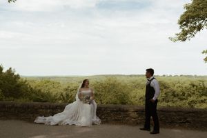 Bride in flowing white gown sits on stone wall as groom stands facing her with scenic green landscape.