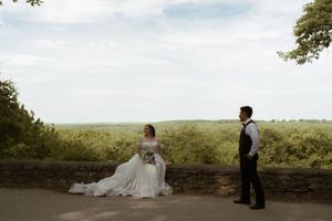 Bride in white gown and groom in black vest share a romantic moment on a stone overlook with scenic views.
