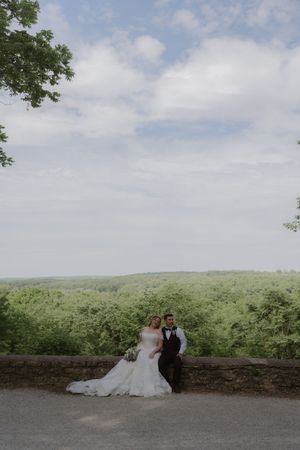 Bride and groom pose against a stone wall with a stunning panoramic green forest landscape backdrop.