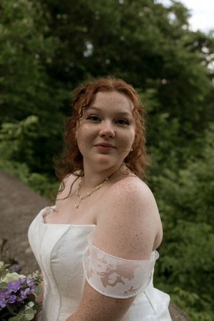 Redhead bride with freckles in off-shoulder white lace wedding dress holding floral bouquet outdoors.