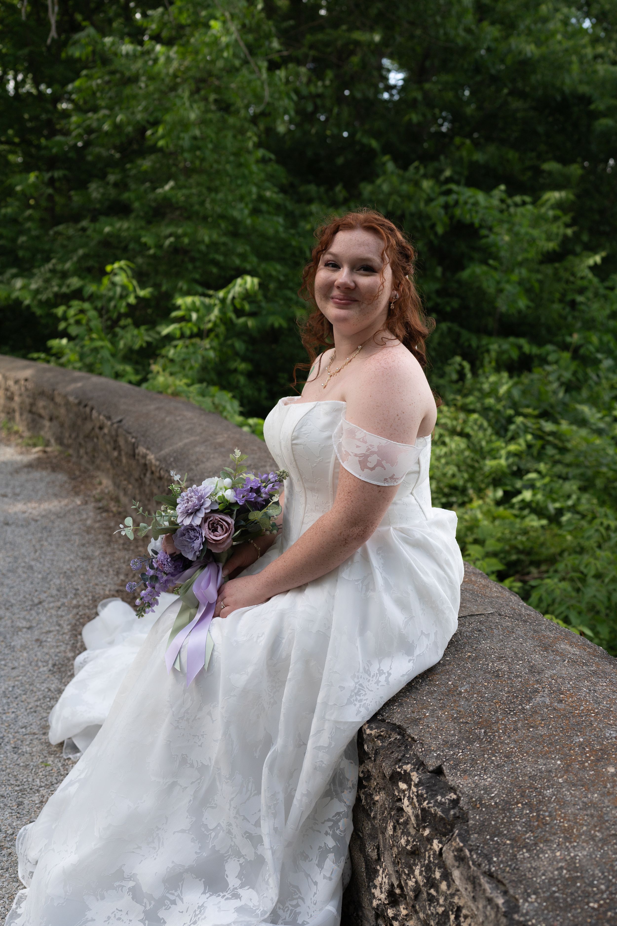 Bride in off-shoulder white gown holding purple bouquet, seated on stone wall surrounded by lush greenery.