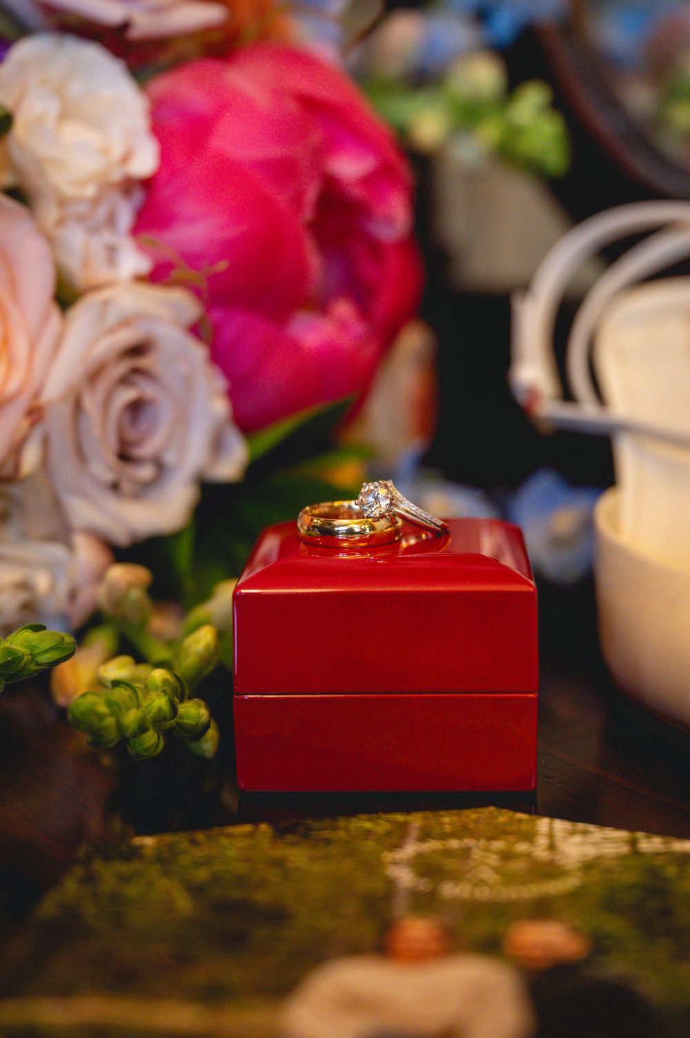 Gold diamond engagement ring resting on a red jewelry box surrounded by pink peonies and roses.
