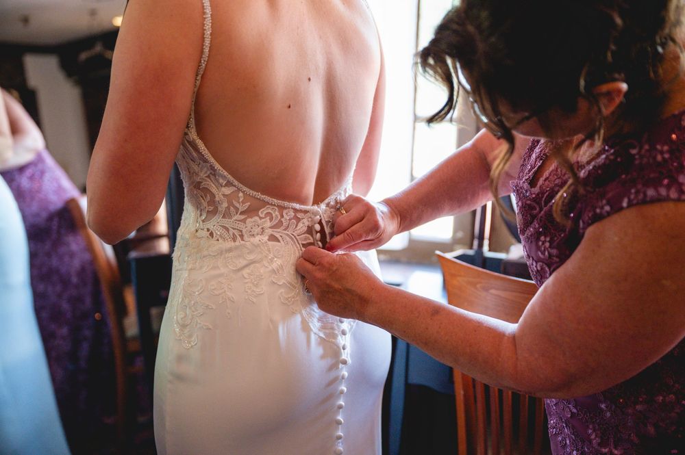 A woman in a white lace wedding gown has her dress buttoned up by a helper in a purple sequined outfit.