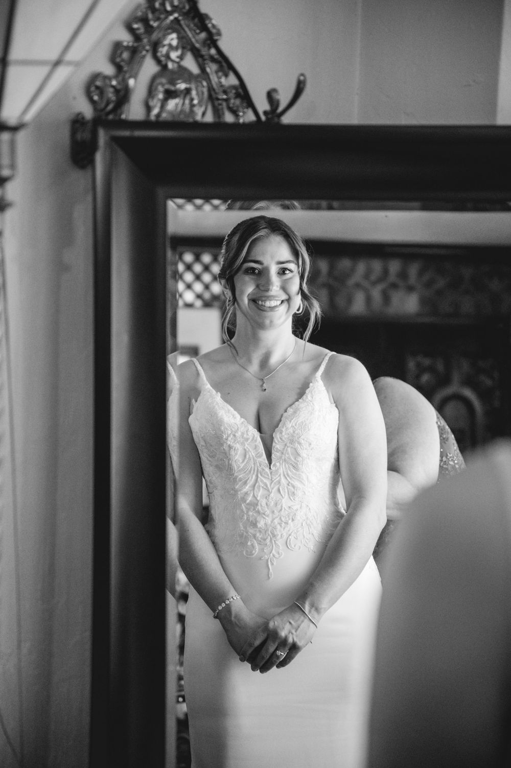 Smiling bride in lace wedding gown stands before ornate mirror in black and white portrait photo.