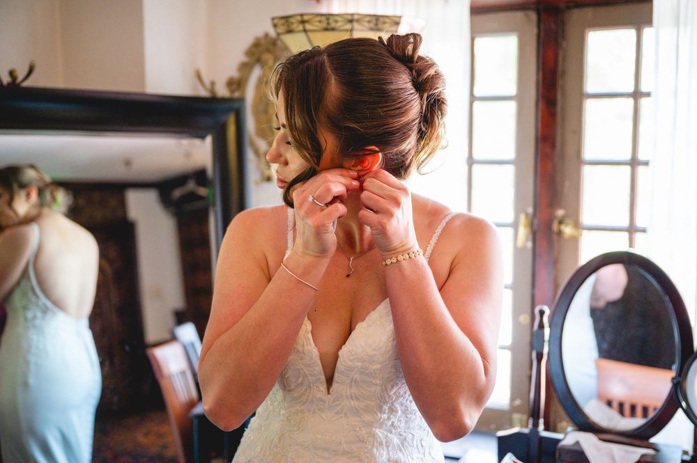 Bride in white wedding dress putting on earrings while getting ready, with mirror reflection in background.