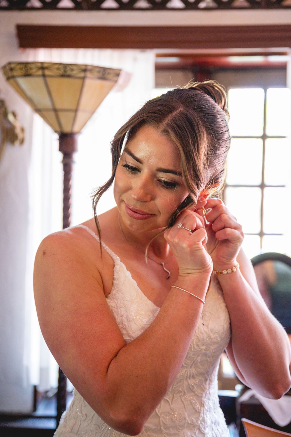 Bride in white lace gown putting on earrings while getting ready, with soft natural light in background.