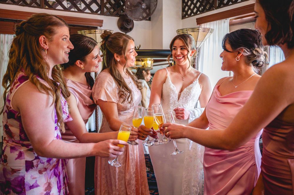 Bride and bridesmaids in pink dresses toasting with champagne glasses during wedding preparations.