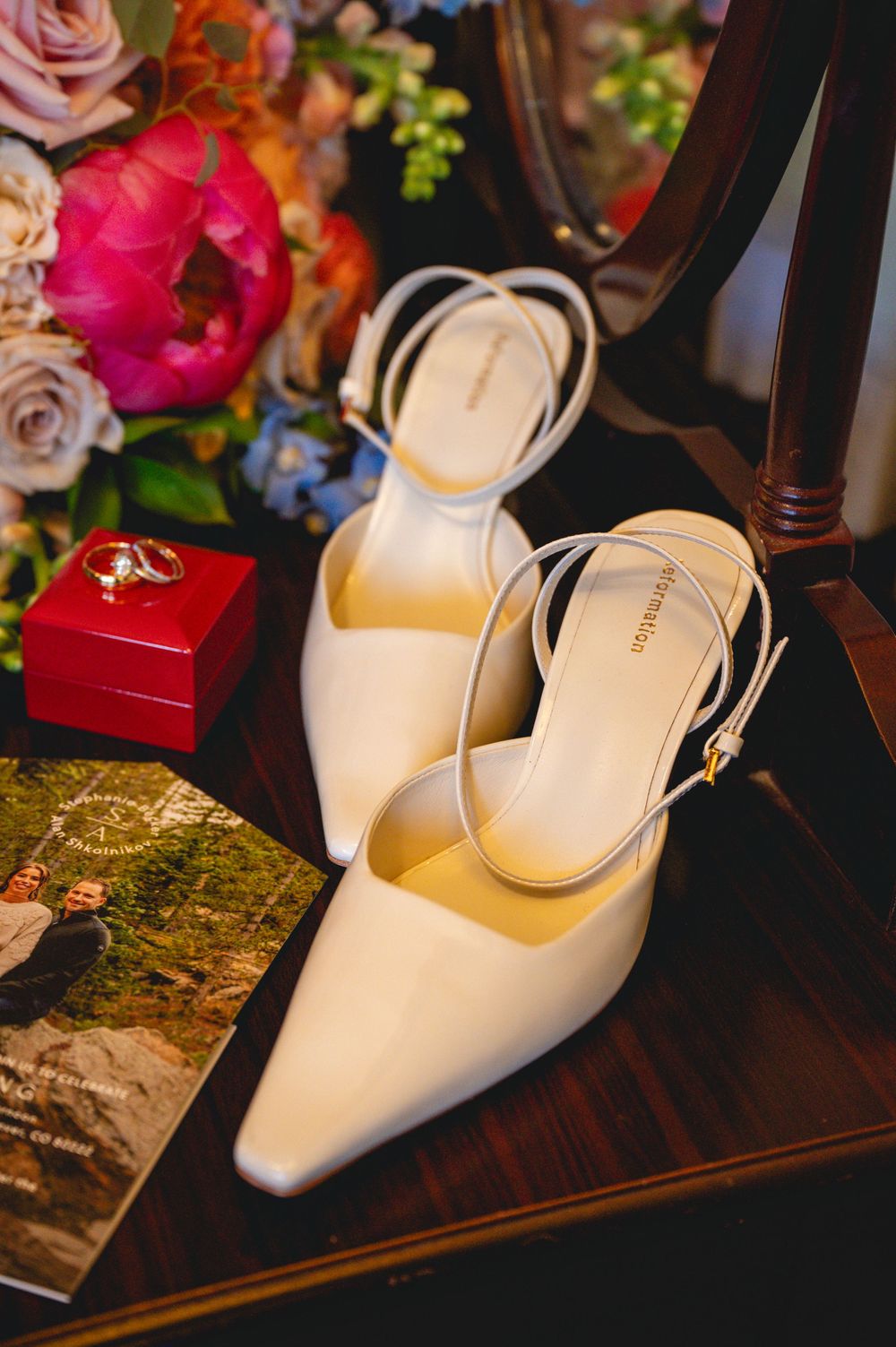 Elegant white pointed-toe kitten heel slingback shoes displayed on a wooden dresser with flowers and a red ring box.