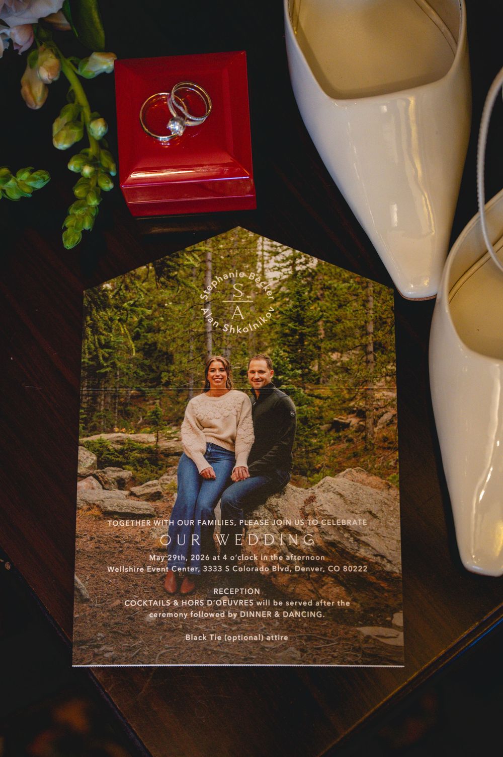 Elegant wedding invitation with couple photo, rings in red box, and white heels on dark wood surface.