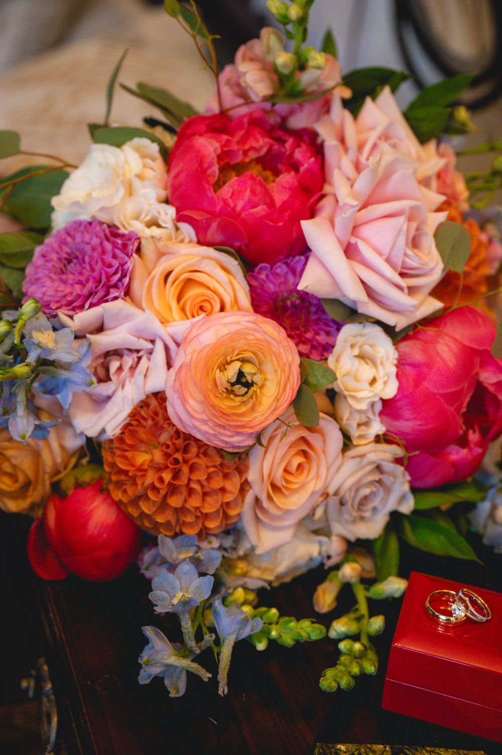 Colorful wedding bouquet with pink peonies, peach roses, orange ranunculus, and blue flowers beside gold rings in a red box.