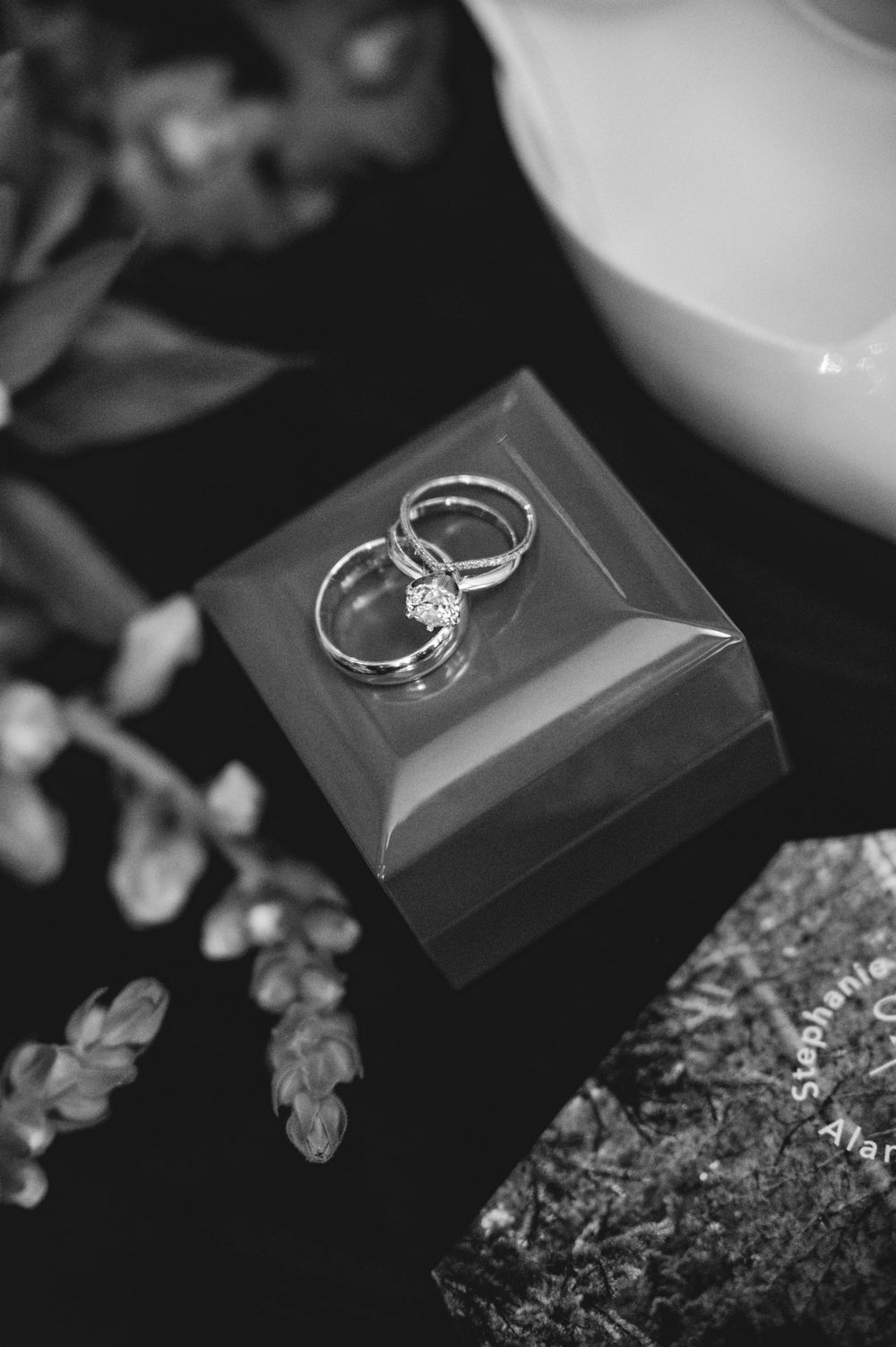Black and white photo of wedding rings displayed on a small jewelry box surrounded by floral decorations.
