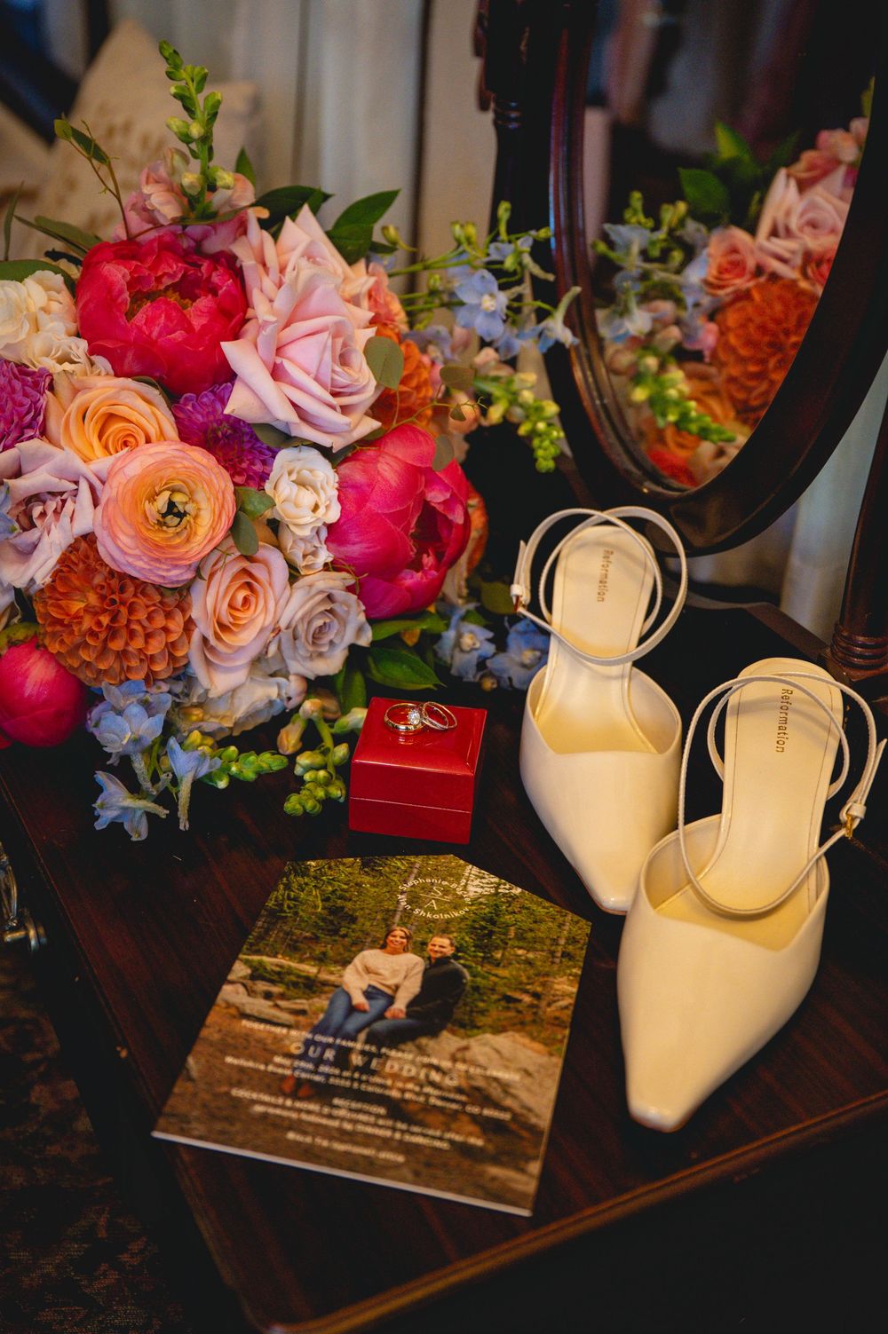 Bridal flat lay with white heels, colorful bouquet of peonies and roses, red ring box, and wedding program on wooden table.
