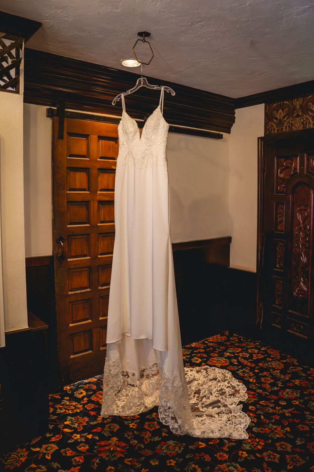 White lace wedding gown hanging from ceiling in elegant wood-paneled room with ornate floral carpet.