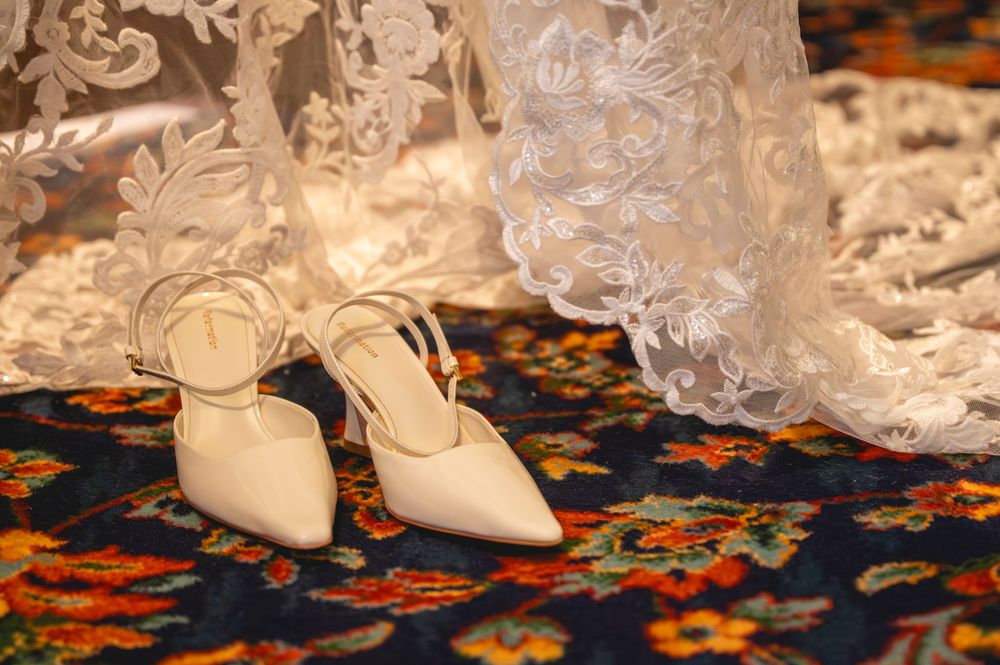 White satin bridal heels with ankle straps rest on a colorful floral carpet beside an elegant lace wedding gown.