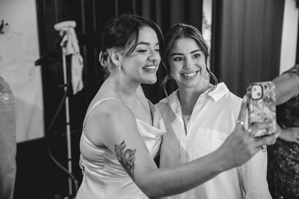 Two women smile while taking a selfie together, one wearing a wedding dress with a tattoo visible on her arm.