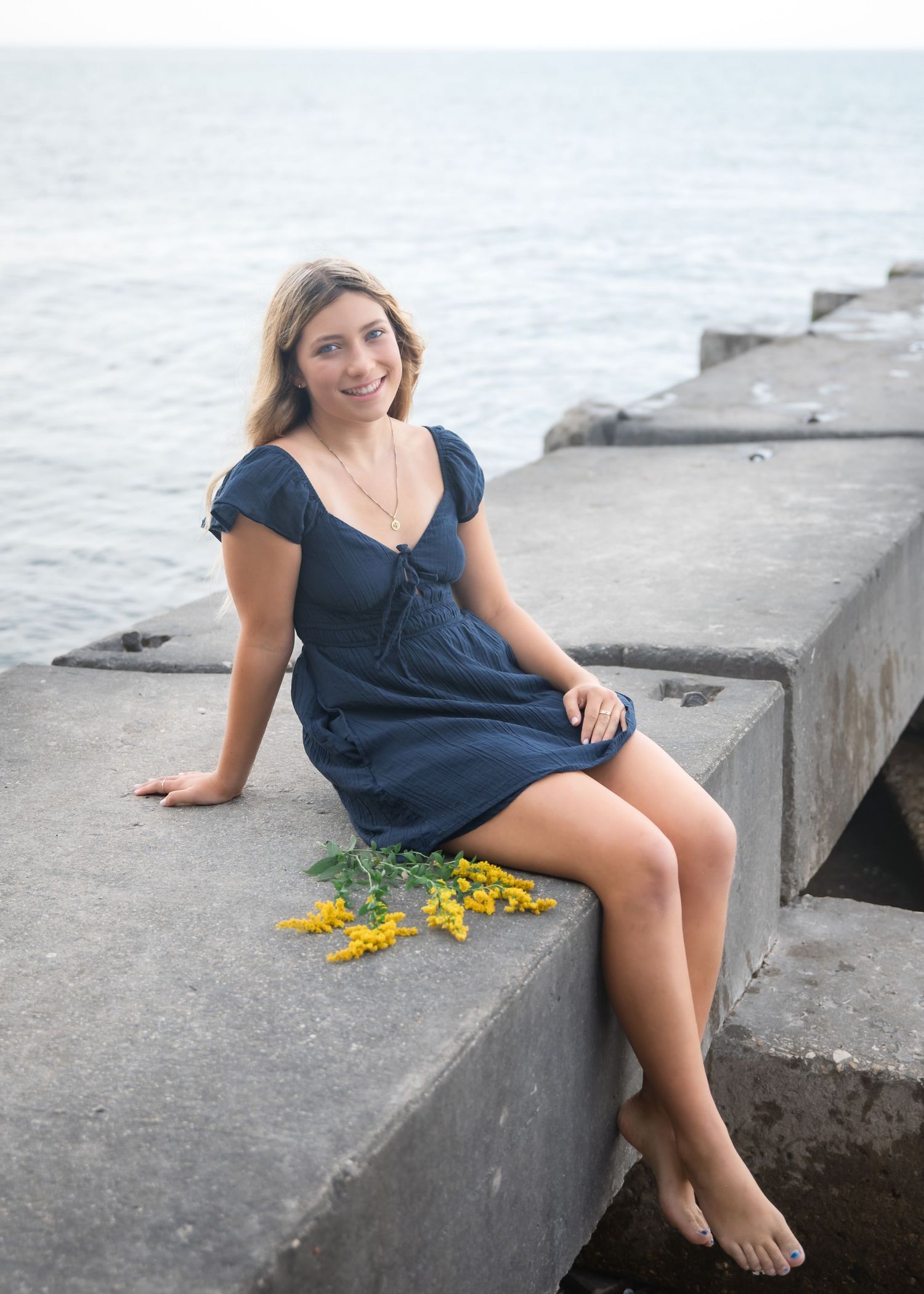 Young woman in navy blue dress sitting barefoot on concrete pier with yellow flowers, smiling with ocean in background.