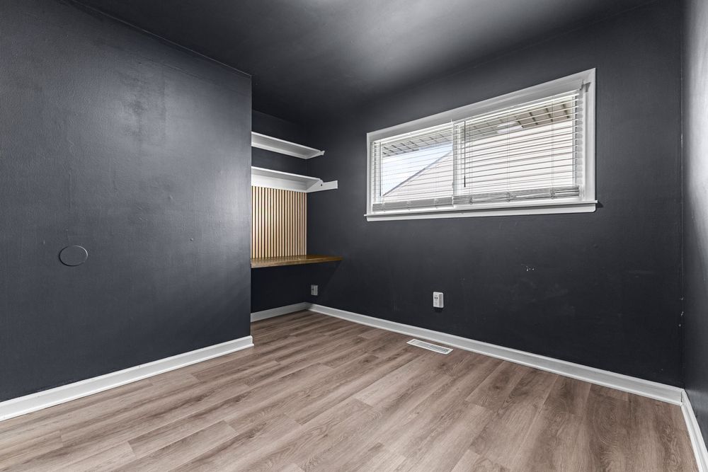 Dark navy bedroom with built-in wooden desk, open shelving, and light hardwood floors with a small window.