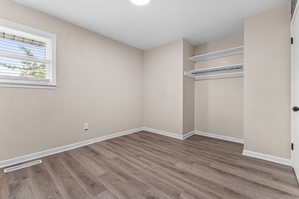 Bright empty bedroom with open closet shelving, light hardwood floors, and a window with natural light.