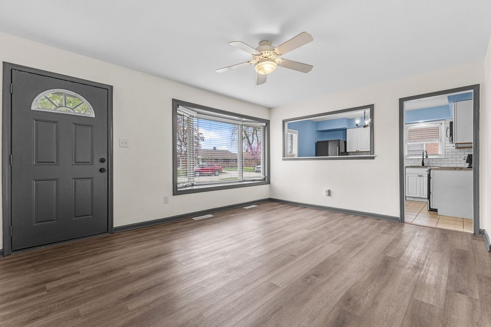 Bright living room with dark hardwood floors, ceiling fan, gray front door with transom window, and open layout.