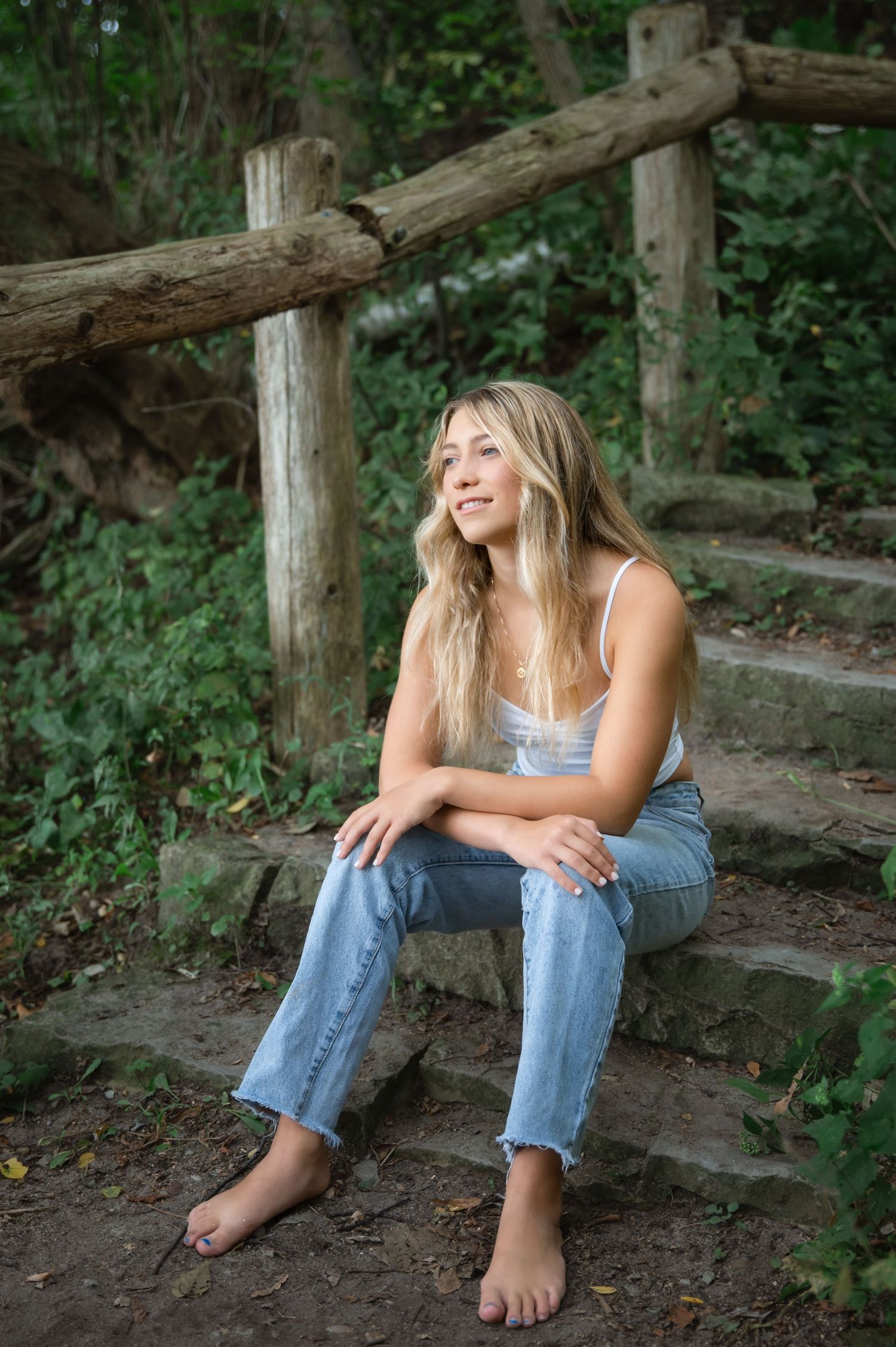 Young woman with long blonde hair sits barefoot on rustic wooden steps in a lush green forest, wearing jeans and a white top.