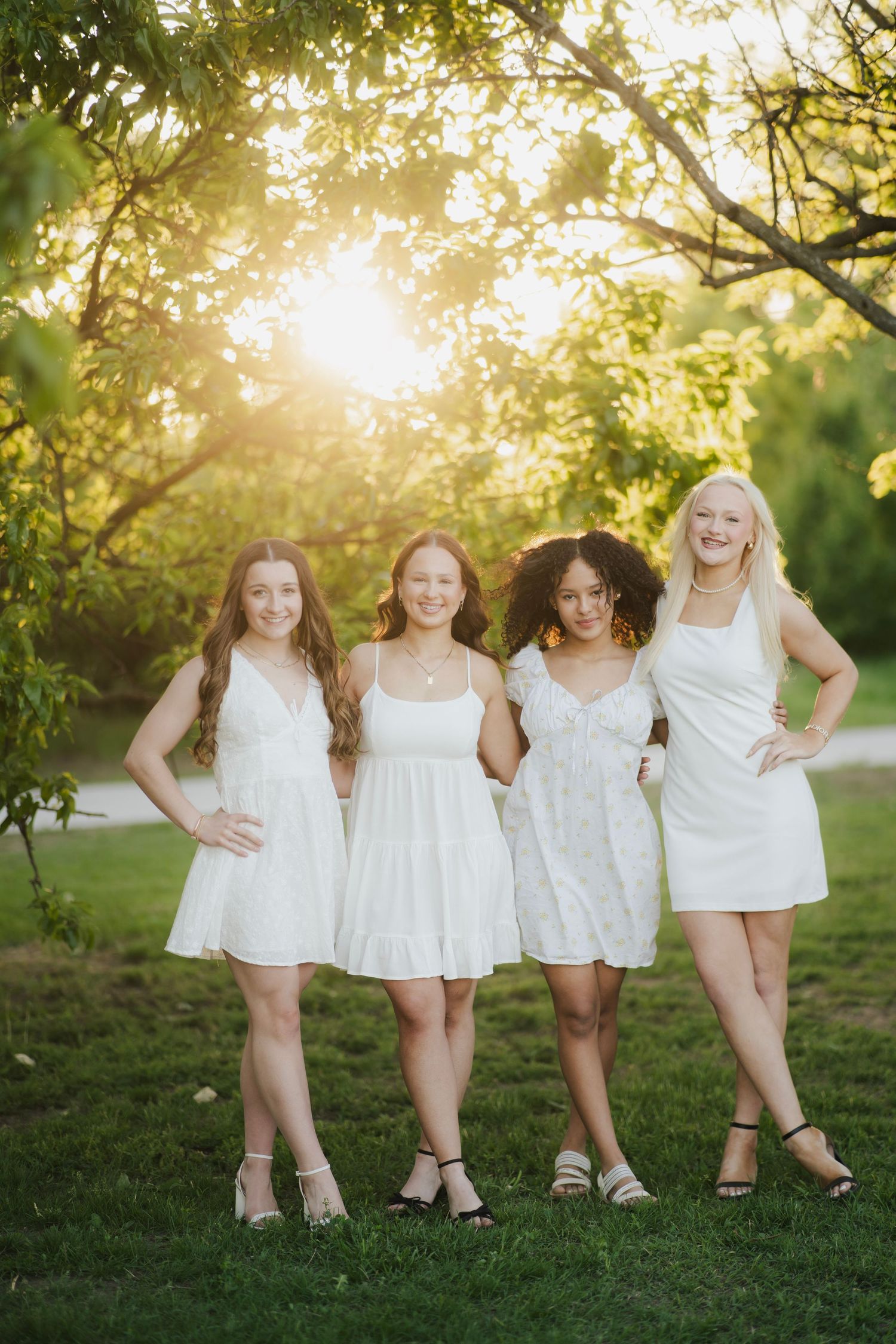 Four young women in white dresses smile together outdoors in warm golden sunset light beneath green trees.